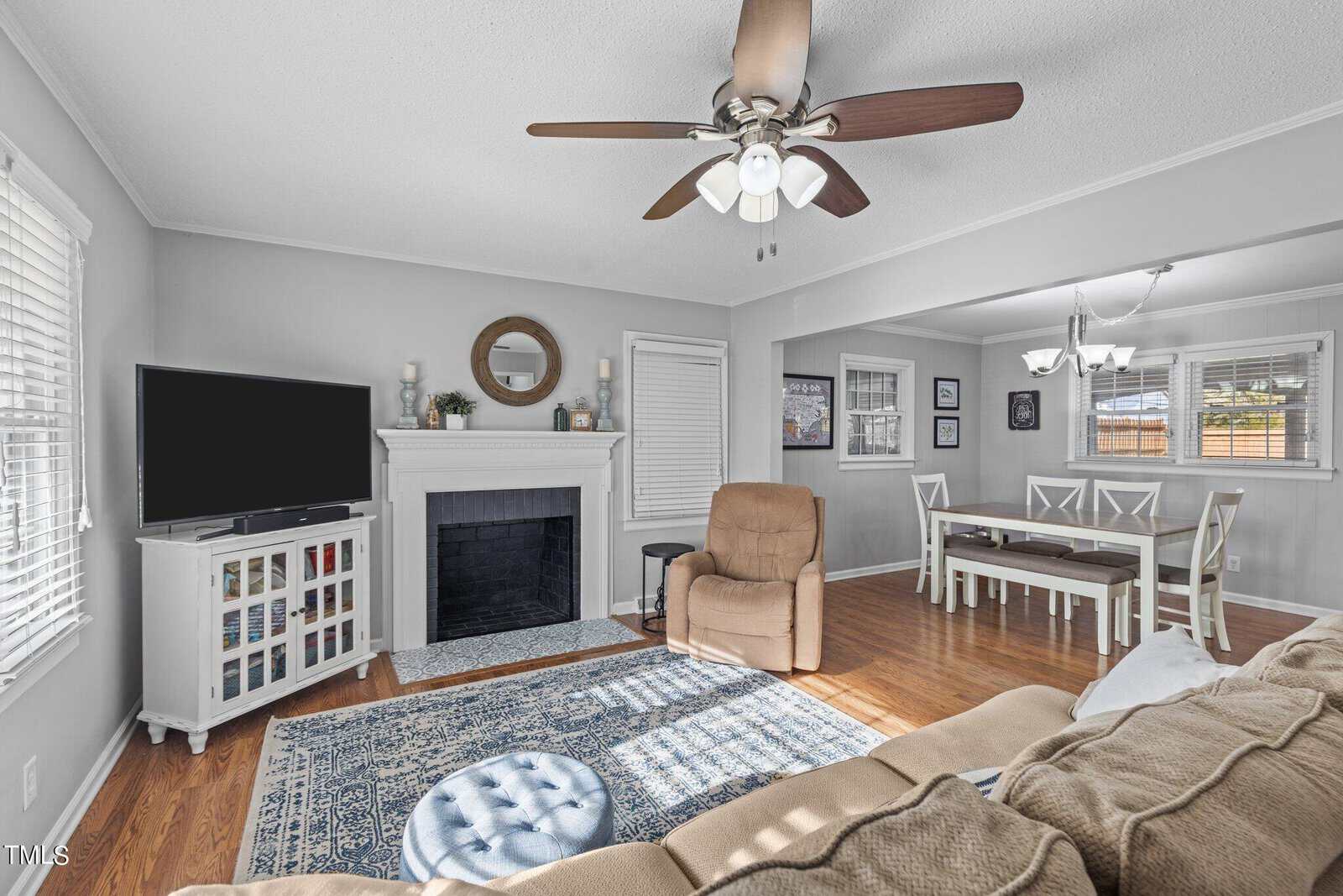 A cozy living room features a beige couch, blue patterned rug, and a wooden coffee table. A recliner sits by a white fireplace with candles and a round mirror. A flat-screen TV is on a stand, and a dining area with a table and chairs is visible.