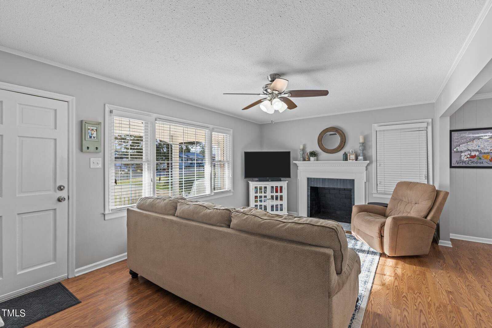 A cozy living room with beige walls features a brown sofa, a recliner, and a fireplace with a mirror above it. A television sits next to the fireplace. The room has wooden floors and large windows allowing natural light to fill the space.