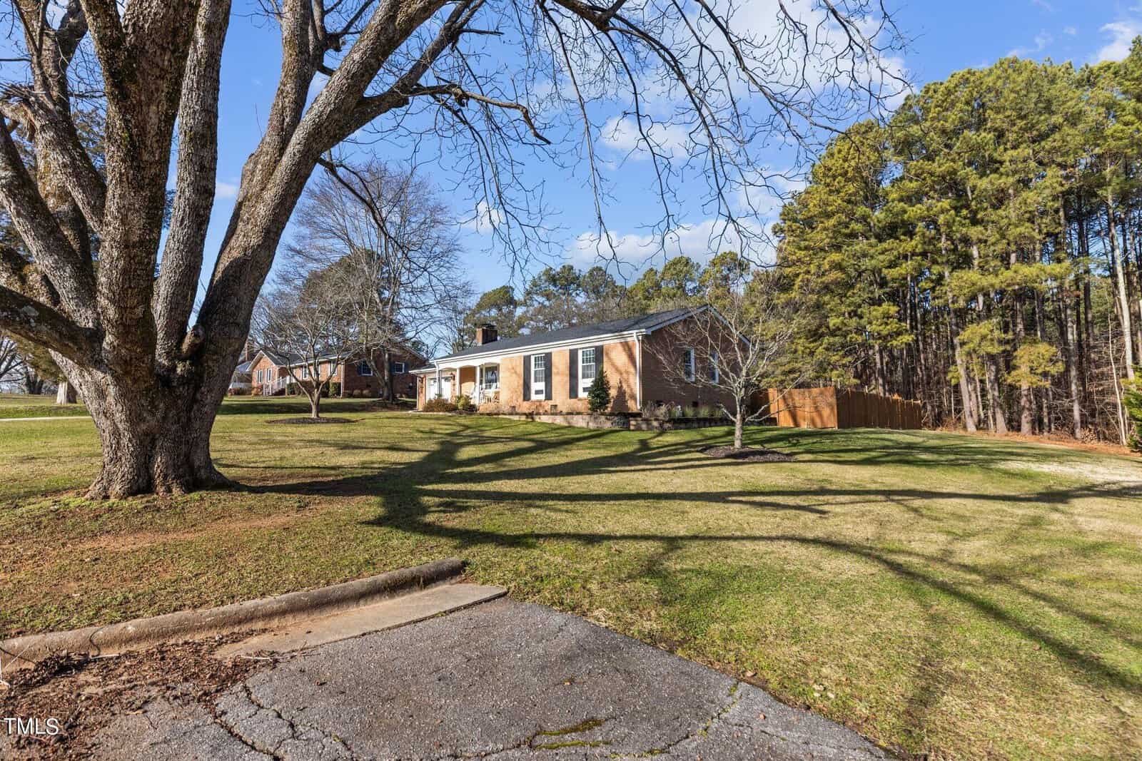 A brick house sits atop a sloping grassy hill with a large tree and a driveway in the foreground. The sky is blue with scattered clouds, and a line of trees borders the property on the right side.