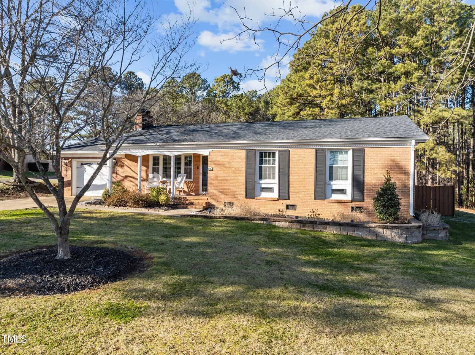 A single-story brick house with a gray shingled roof, three front windows with white shutters, and a small front porch. The house is surrounded by a grassy yard and tall trees in the background under a blue sky.