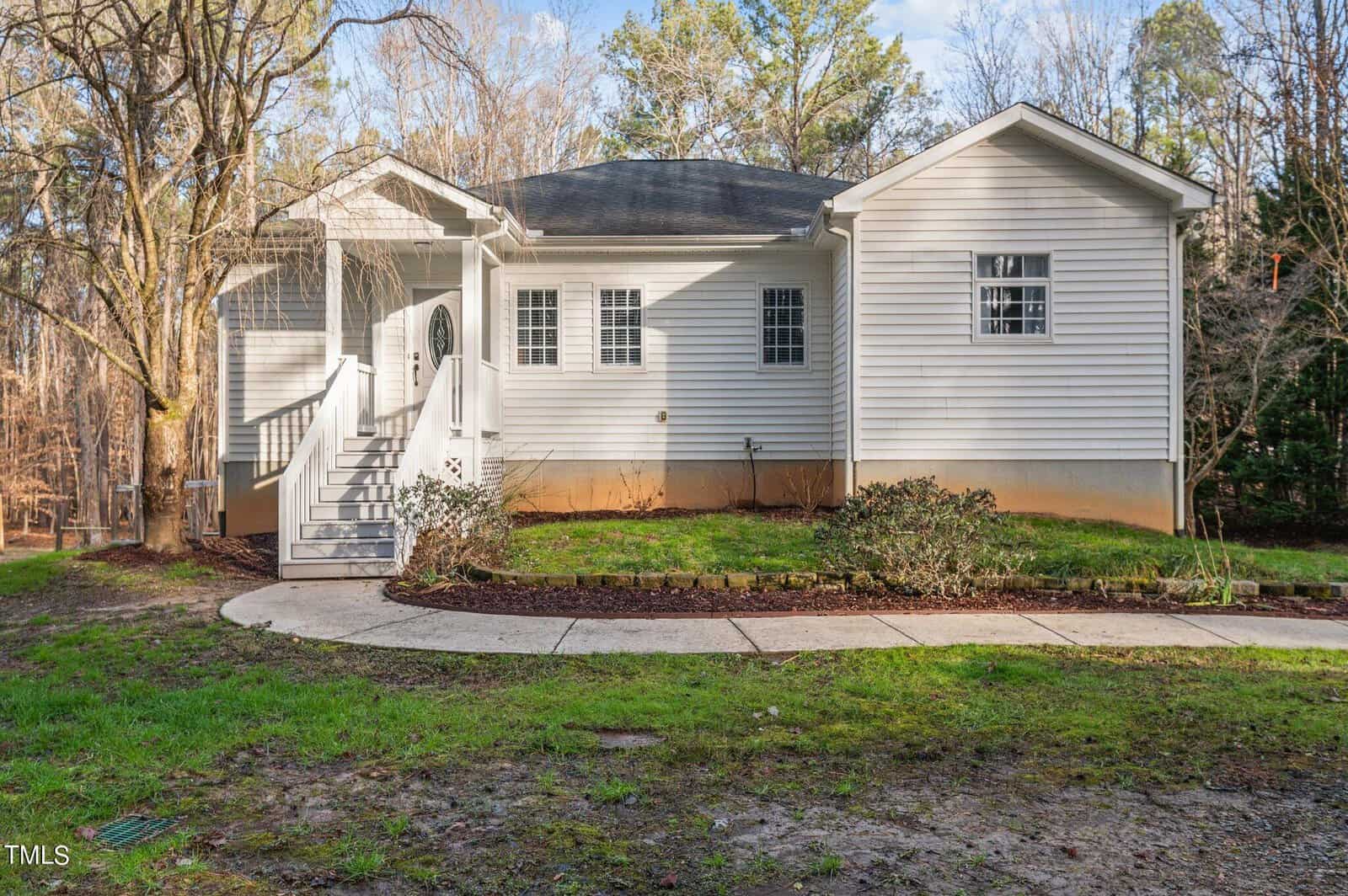A small white house with a gabled roof surrounded by trees. It features a front porch with steps leading up, white siding, and several windows. A curved sidewalk and a grassy lawn are in the foreground.