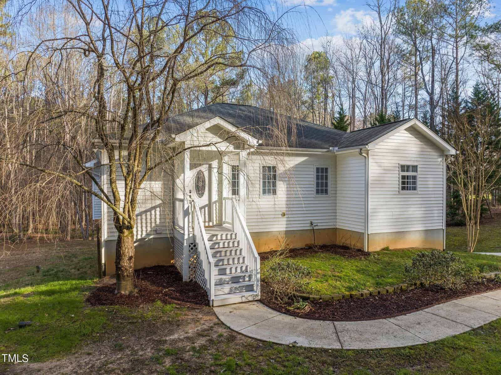 A small white house with a gabled roof is surrounded by trees. The entrance features a white staircase leading up to a porch with a circular window in the front door. The yard is grassy with a concrete walkway.