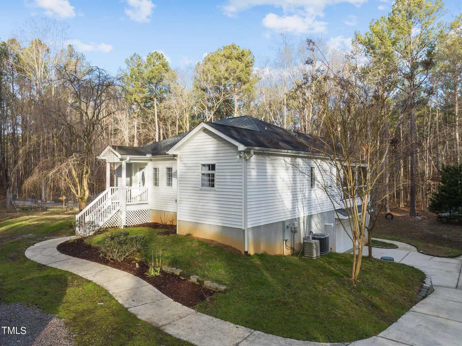 A small white single-story house with a raised porch and railing is surrounded by a wooded area. The property has a curved paved driveway and a trimmed lawn under a clear blue sky. Trees and shrubs are in the background.