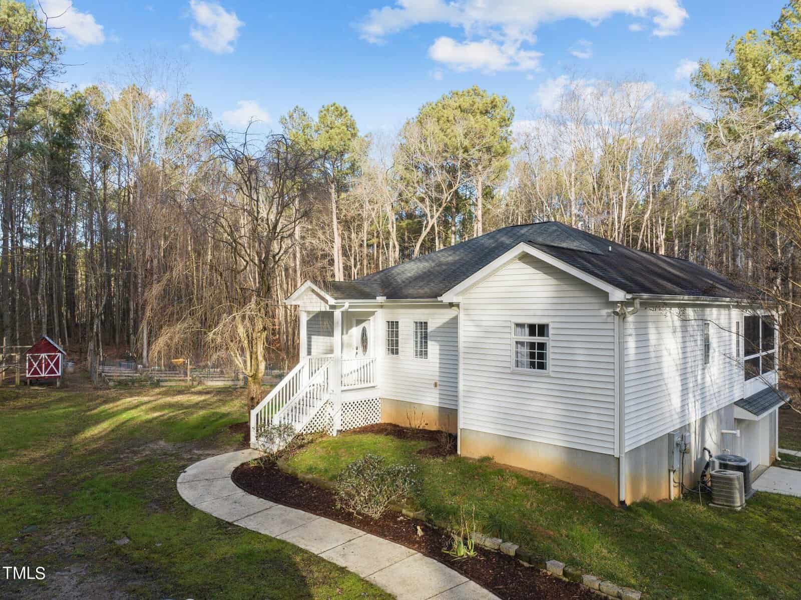 Nestled on 3697 Hugh Davis Road in Youngsville, a white house with a porch and black roof overlooks a yard with a paved walkway. A red barn-like structure stands in the background, bordered by a forest of tall trees under a partly cloudy blue sky.