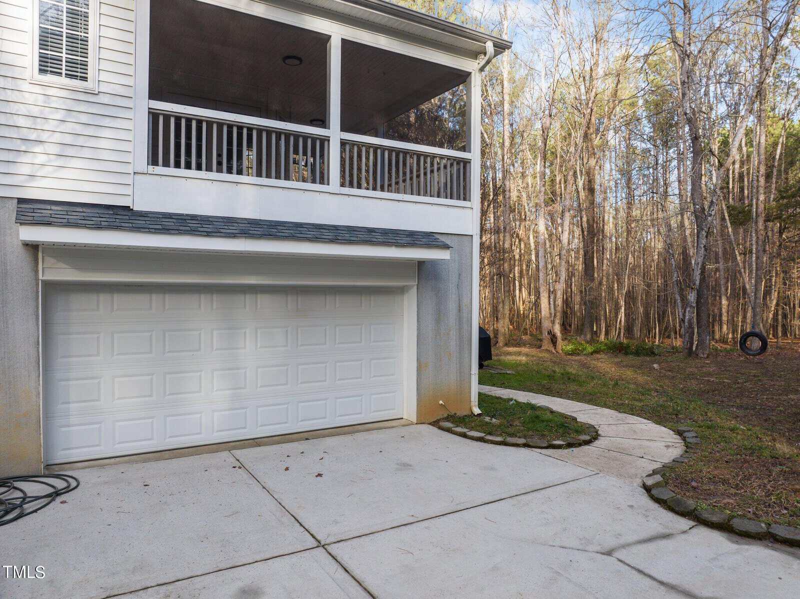 A two-story house with a garage on the lower level and a covered porch above. The driveway is concrete, leading to the garage door. The house is surrounded by trees, and a small stone-bordered patch of grass is visible to the side.