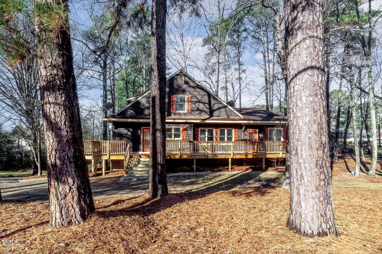 A rustic, dark brown, two-story wooden cabin with a large front porch is surrounded by tall pine trees and a forest floor covered in pine needles. The sky is partly cloudy, casting soft light on the scene.