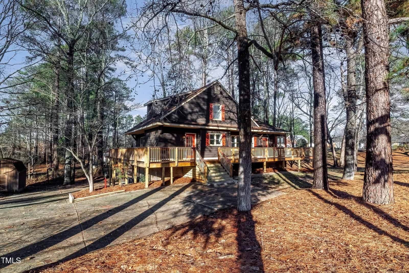 A rustic wooden cabin surrounded by tall pine trees in a forest setting. The cabin features a large wraparound deck and is set on a sloped, pine needle-covered ground. The sky above is clear and blue, suggesting a sunny day.