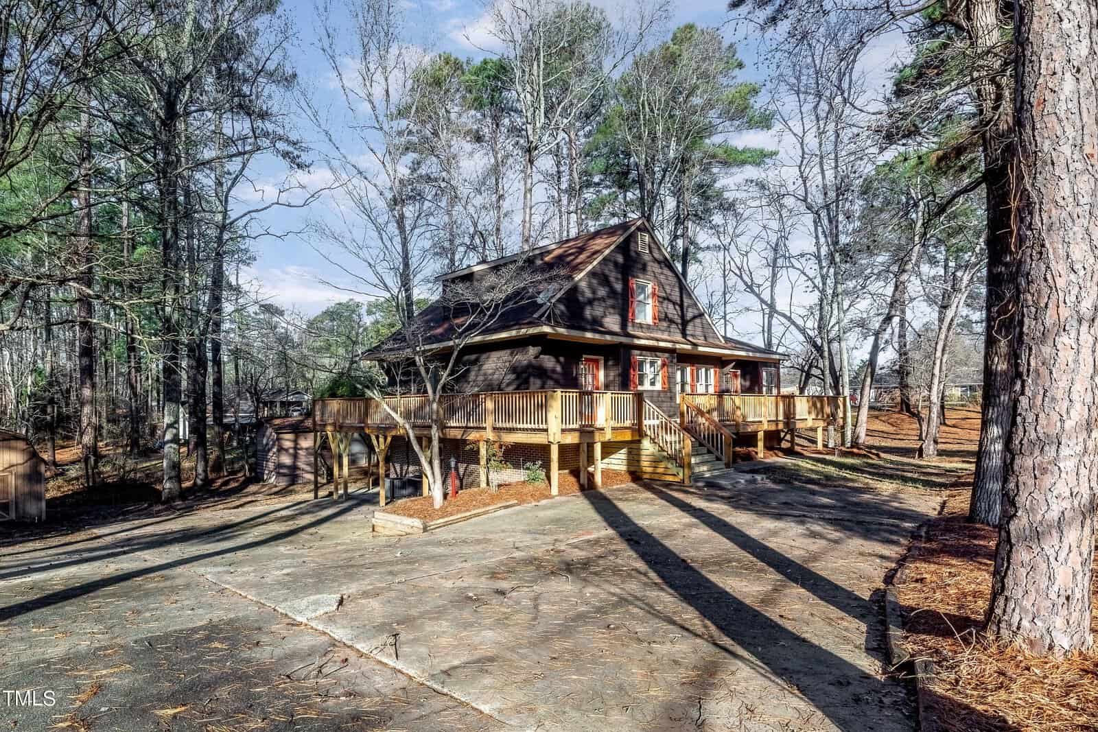 A rustic cabin-style house with a wooden deck is surrounded by tall trees. The driveway leads up to the entrance, and the house features a steep, dark roof and several windows. The setting is wooded and peaceful.