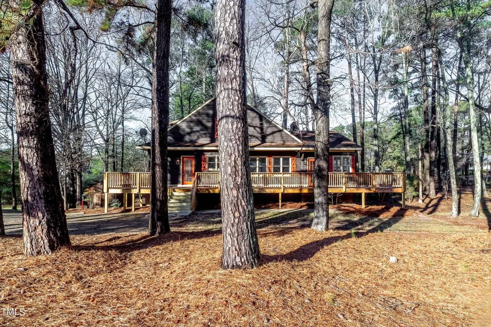 A rustic cabin surrounded by tall pine trees with a large wooden deck. The ground is covered in pine needles, and the trees' shadows stretch across the scene, creating a tranquil forest atmosphere.