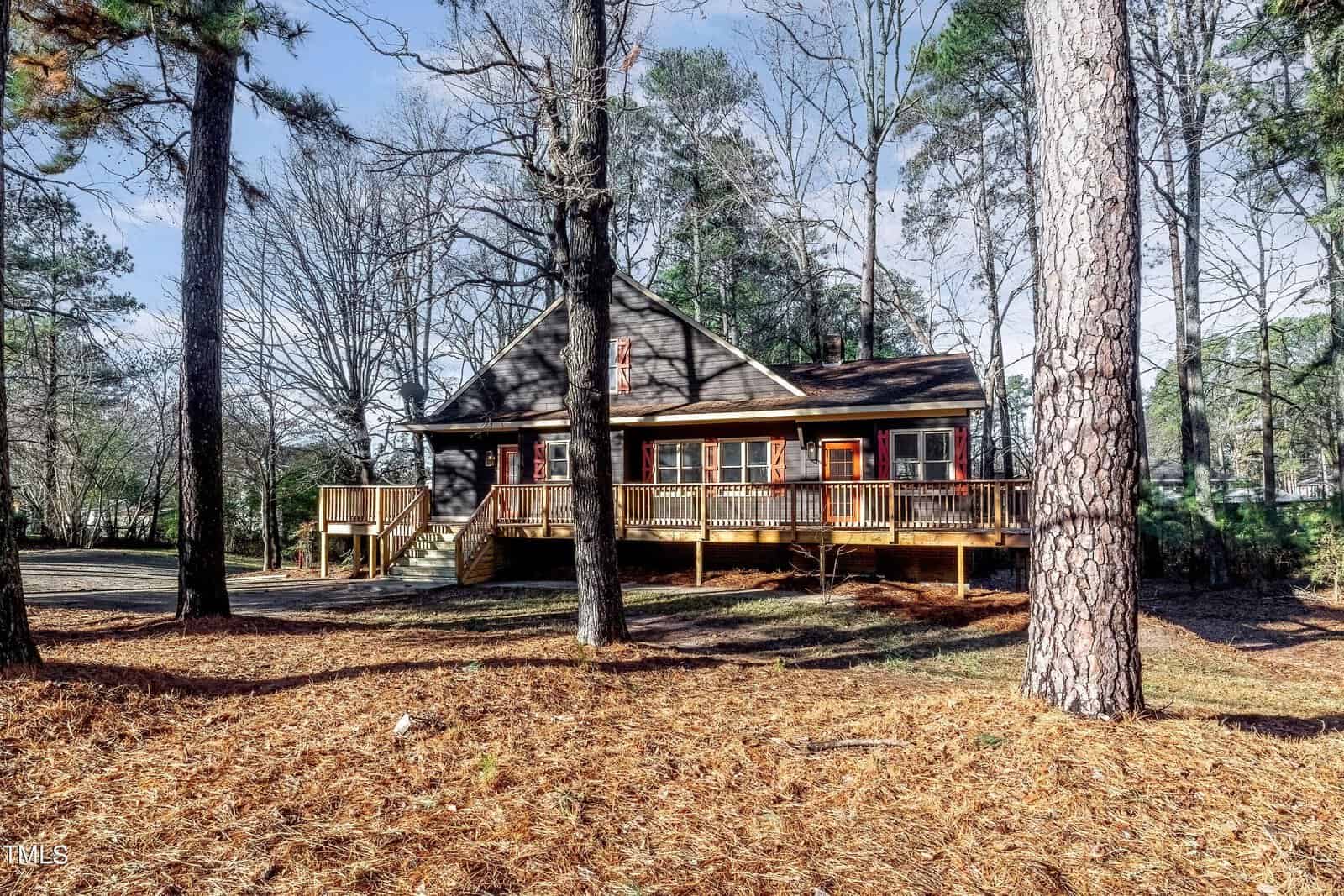 A rustic wooden house with a wraparound porch surrounded by tall pine trees. The yard is covered in brown pine needles and the sky is partly cloudy. The setting conveys a serene and natural ambiance.