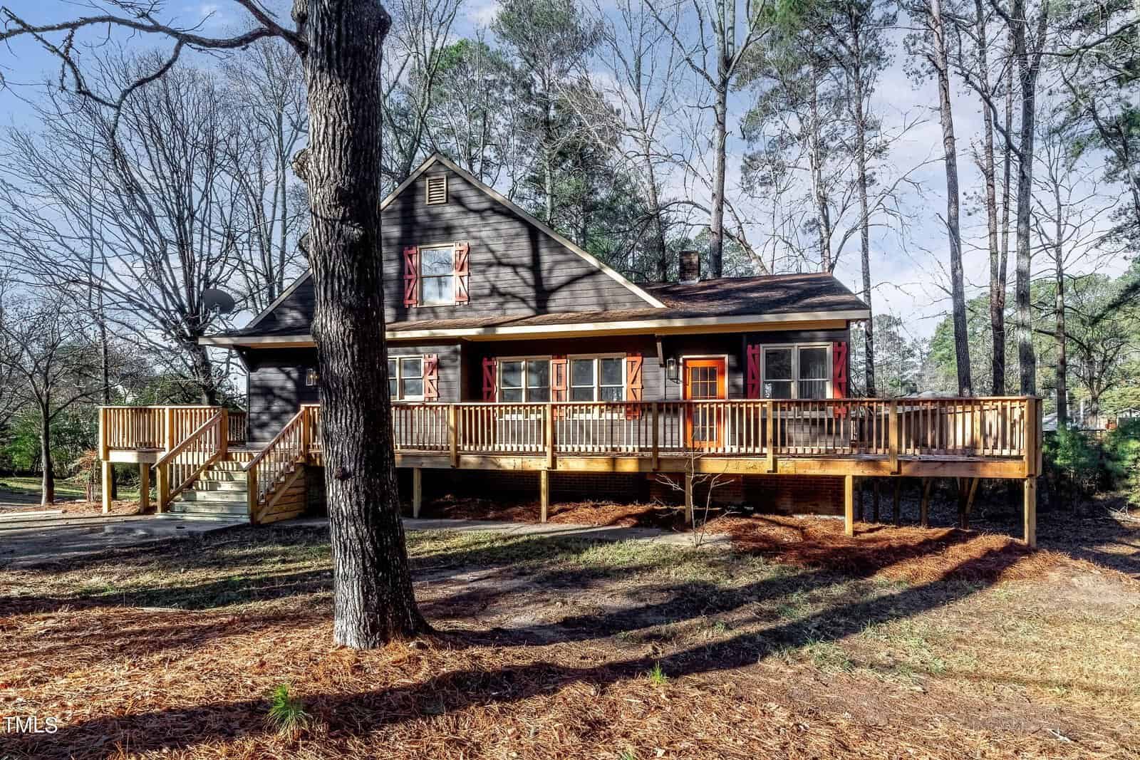 A rustic wooden house surrounded by tall trees and a spacious deck. The exterior features brown siding with red shutters. The yard is covered with pine needles and bathed in sunlight on a clear day.