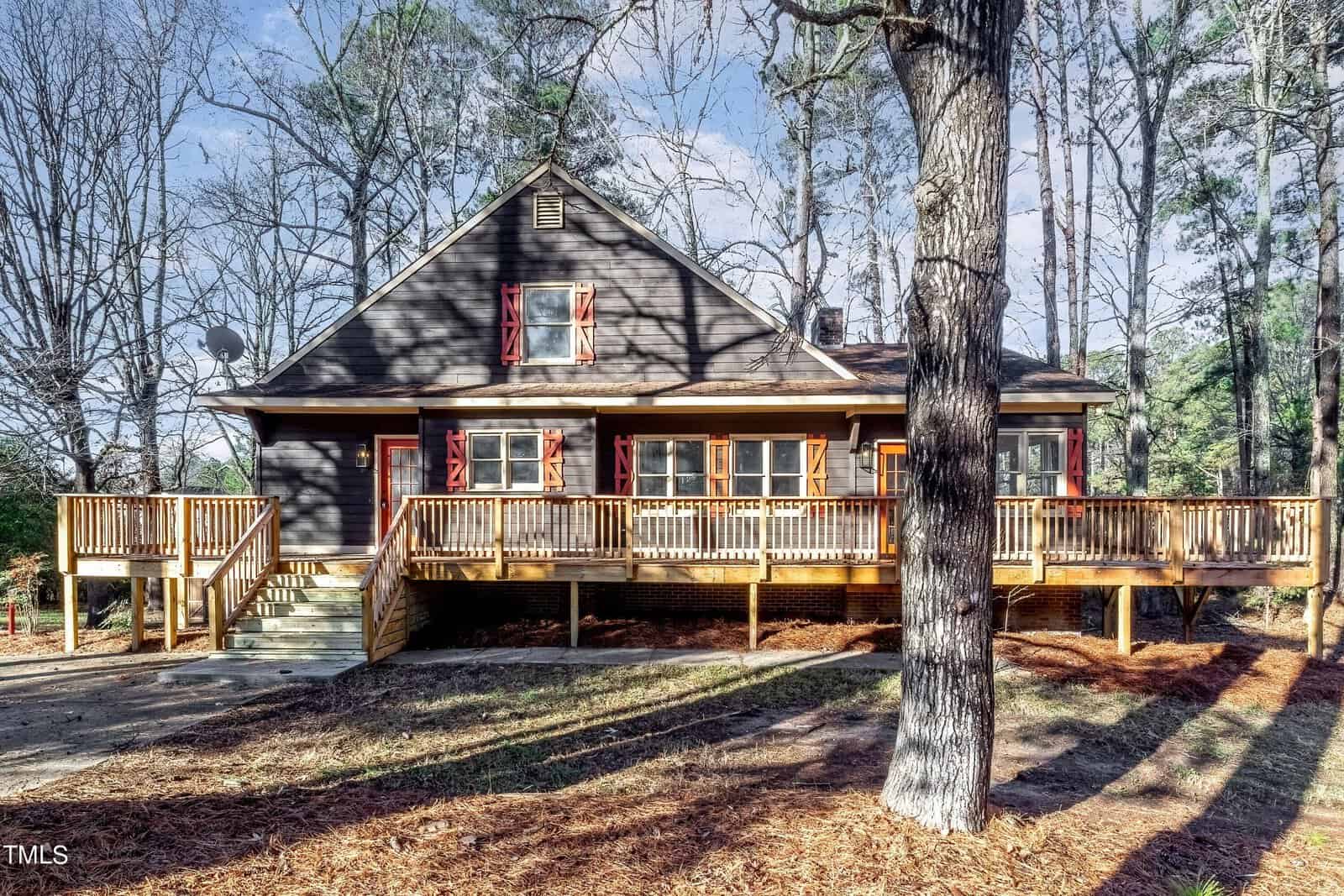 A rustic brown cabin on Creedmoor with a large wooden deck, surrounded by tall trees. The front features red shutters on windows and a wooden staircase leading to the entrance. It's set against a backdrop of bare winter trees and scattered pine needles.