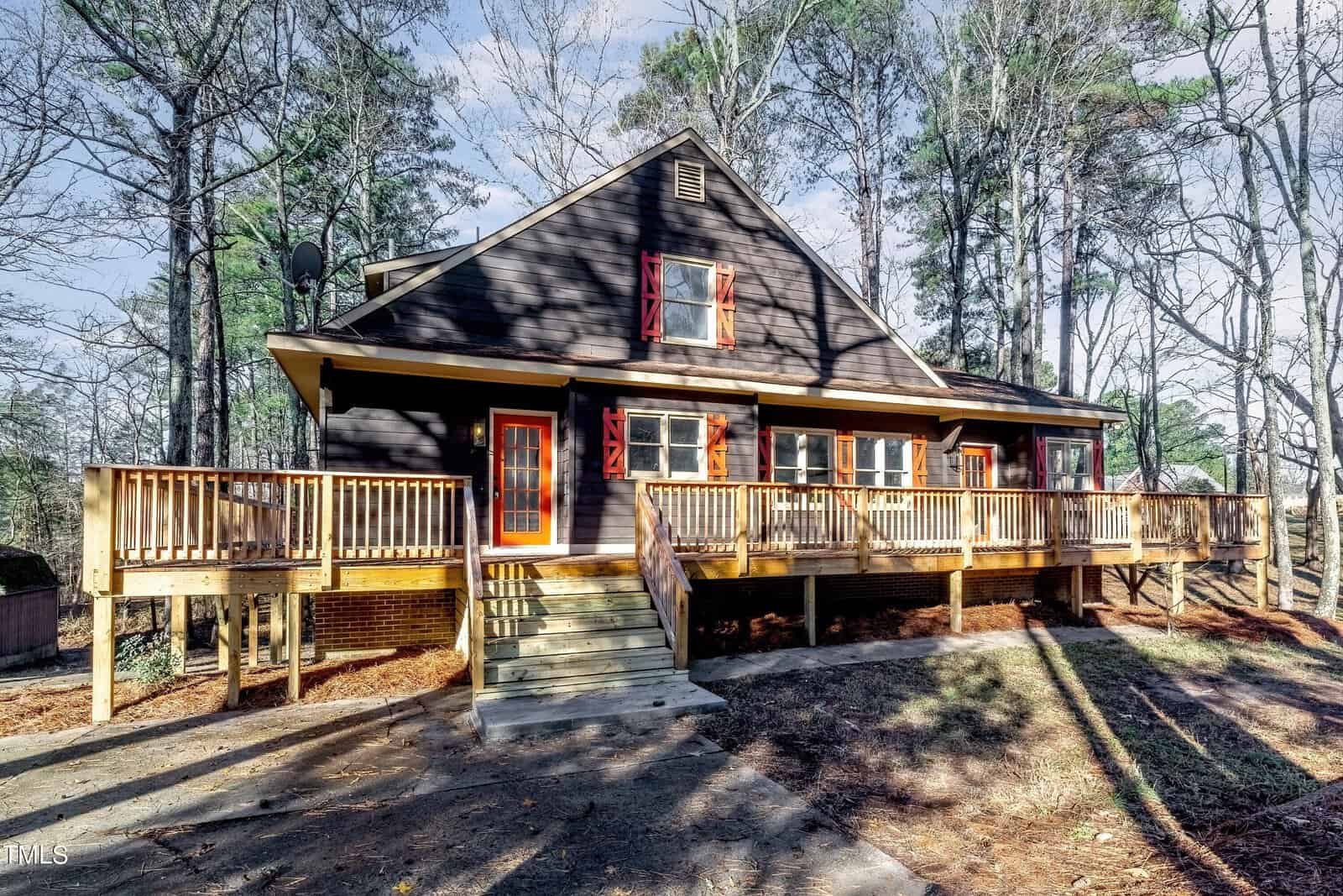A rustic, dark wooden house with red window shutters, surrounded by tall trees. The front features a wide wooden porch with railings and a central staircase leading to the entrance. The setting is serene and wooded, under a clear blue sky.