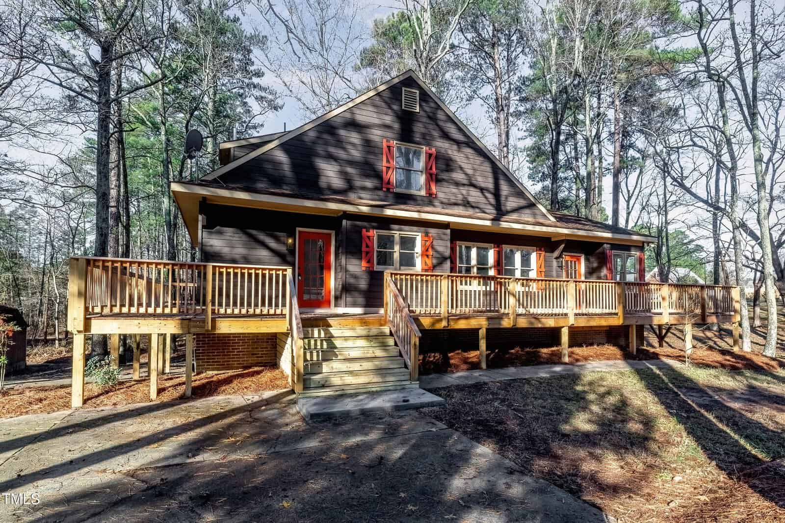 A rustic, two-story cabin with dark wooden siding, red trim, and red shutters is surrounded by tall pine trees. It features a large wooden deck with stairs leading down to a concrete path amidst a wooded area.