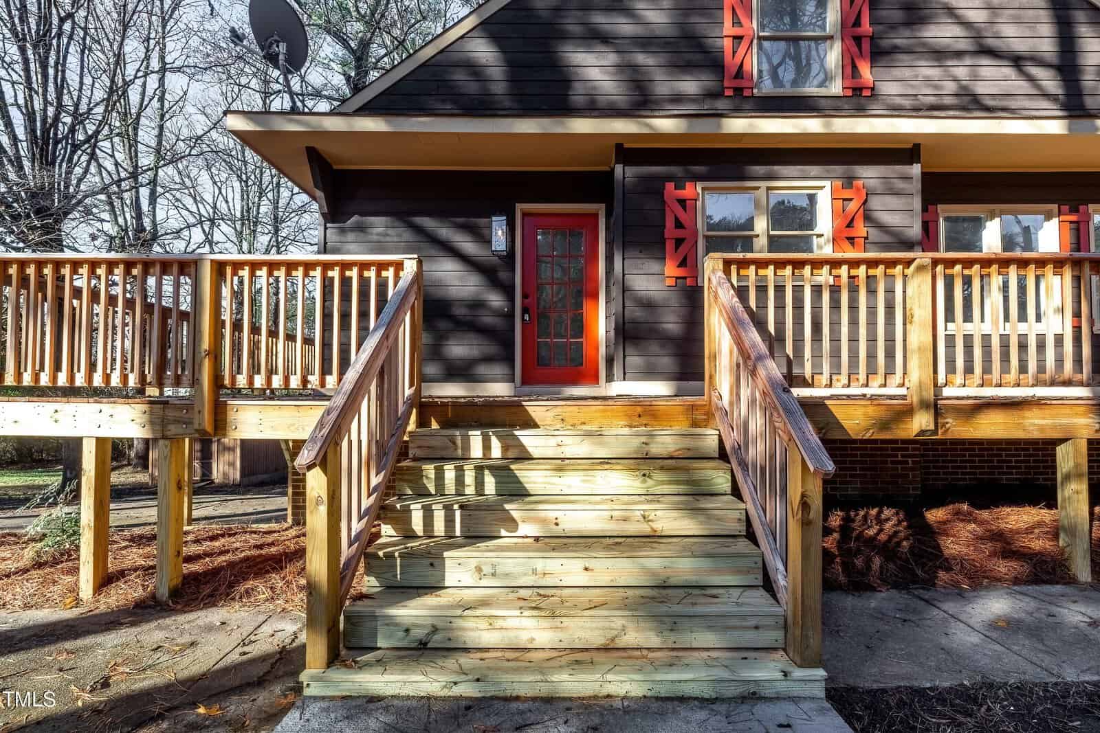 A wooden staircase leads to a house with dark siding and a vibrant red door. The house features red window shutters and a spacious deck. Leafless trees are visible in the background, suggesting a winter setting.