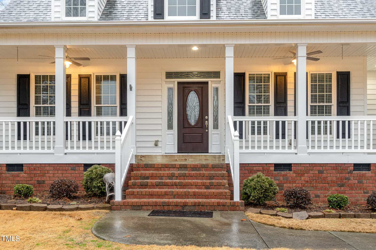 A white house with a covered porch and steps leading to a dark wooden front door. The porch features white railings and columns, with two ceiling fans. The exterior includes brick accents and a shingled roof. Shrubs are planted along the front.