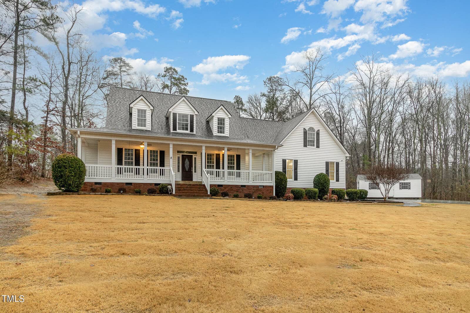 A spacious white house with gray roofing, featuring a large porch and multiple gables, is set against a backdrop of leafless trees under a partly cloudy sky. A manicured lawn stretches in front, and a small white shed is visible to the right.