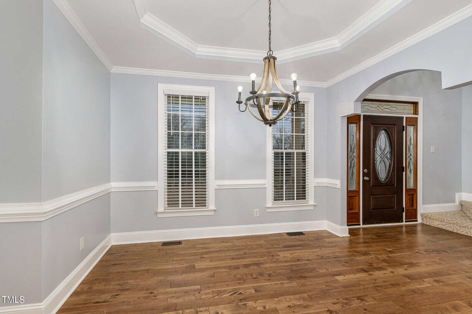 A bright, empty room with light gray walls and a wooden floor. The room features a chandelier, two tall windows with white blinds, and a dark wooden door with decorative glass panels. White trim accents the room, including a tray ceiling and wainscoting.