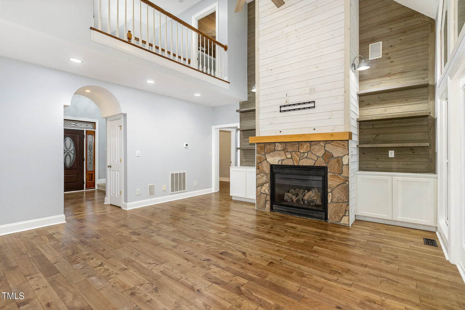 A spacious living room with wooden floors, a high ceiling, and a tall stone fireplace. The walls are light gray, and a wooden staircase with a white railing is visible on the upper level. An arched doorway leads to another room.