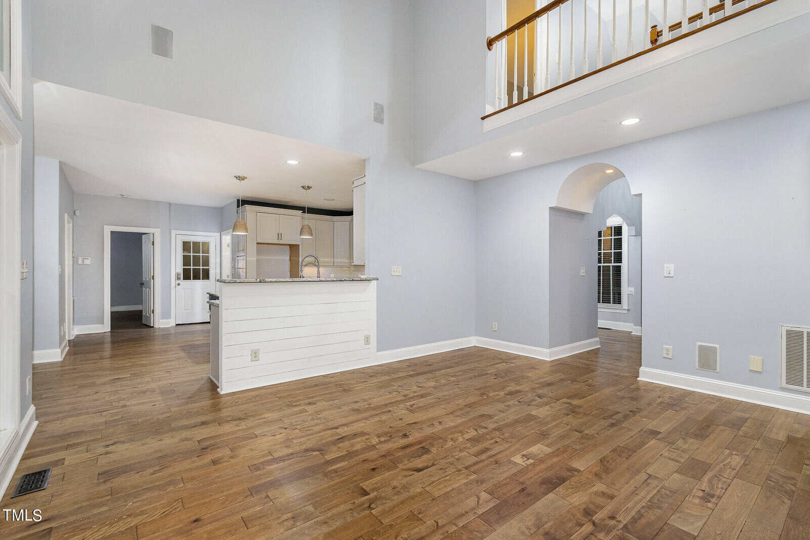 Spacious living area with high ceilings, hardwood floors, and light gray walls. Features include an open kitchen with a white island, recessed lighting, and a railing on the upper level. Arched doorway and a door leading to the backyard are visible.