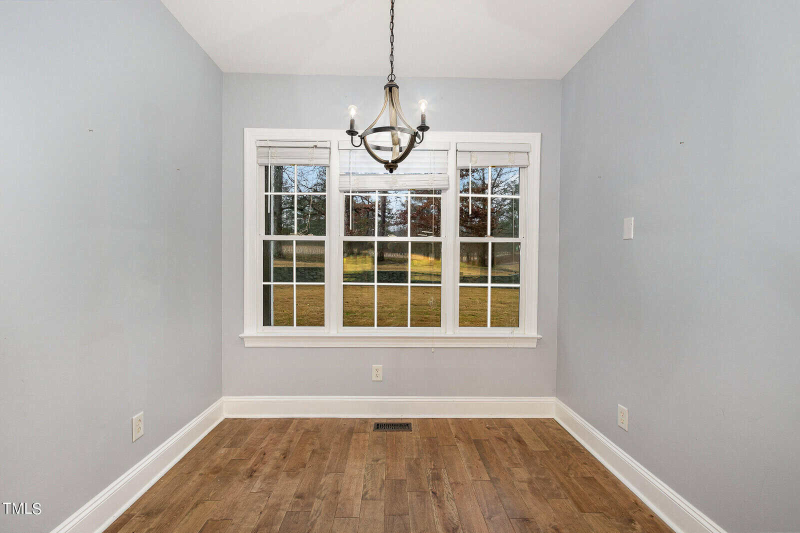 An empty room with gray walls and a wooden floor features a large window with multiple panes and a view of a grassy yard. Above, a modern chandelier hangs from the ceiling.