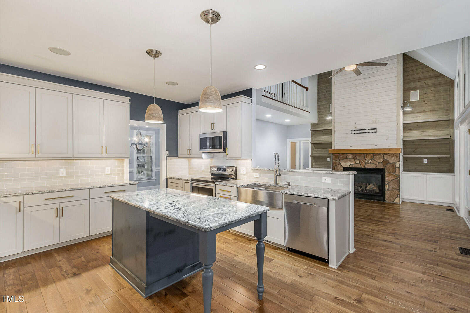 A modern kitchen with white cabinets, a dark island with grey granite countertop, stainless steel appliances, and hardwood floors. An open living space features a stone fireplace and high ceilings with a wooden accent wall.