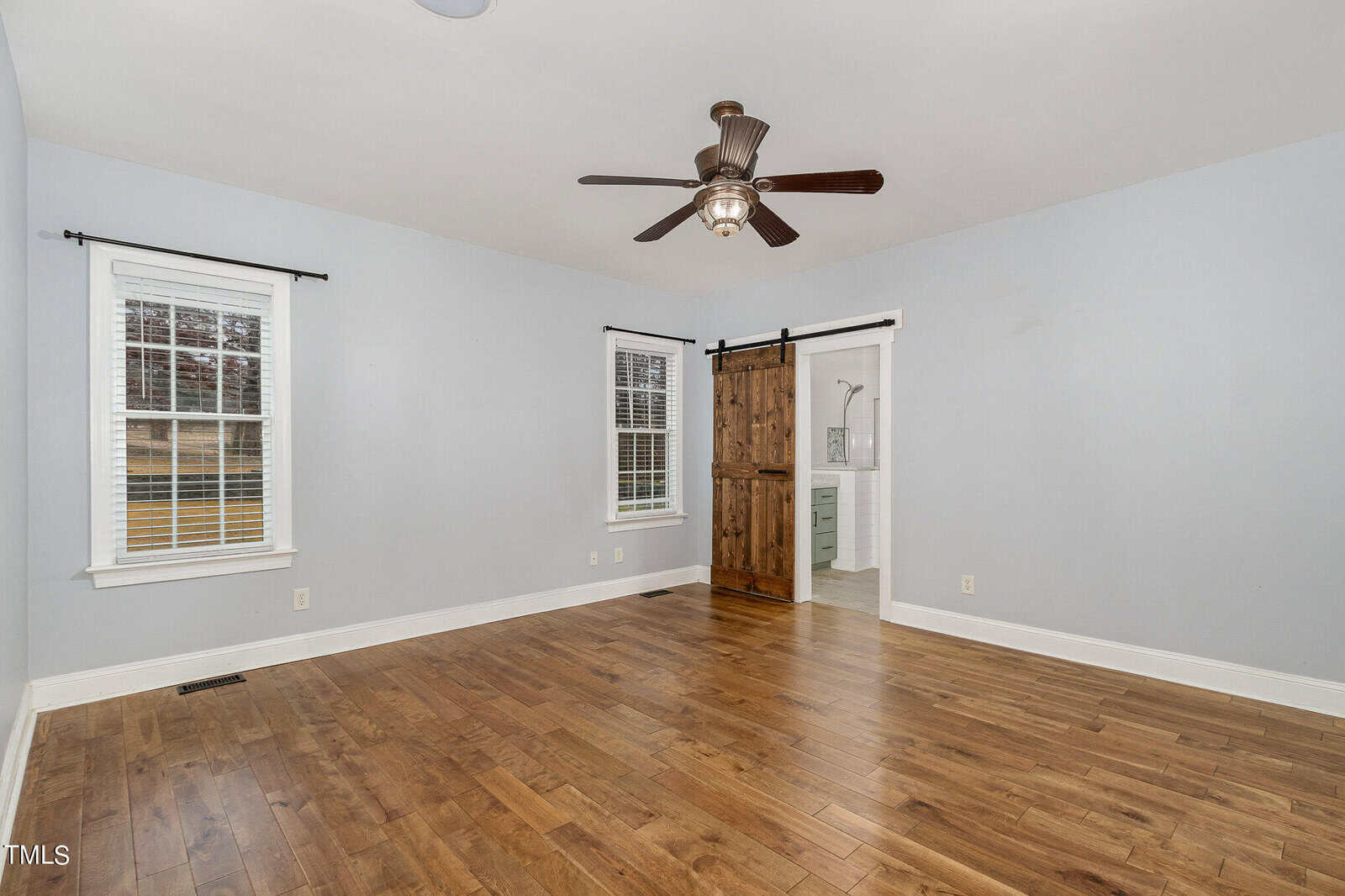 A spacious, empty room with light gray walls and hardwood flooring. Two windows are on the left wall, and a ceiling fan is in the center. A sliding wooden barn door leads to an adjacent bathroom.