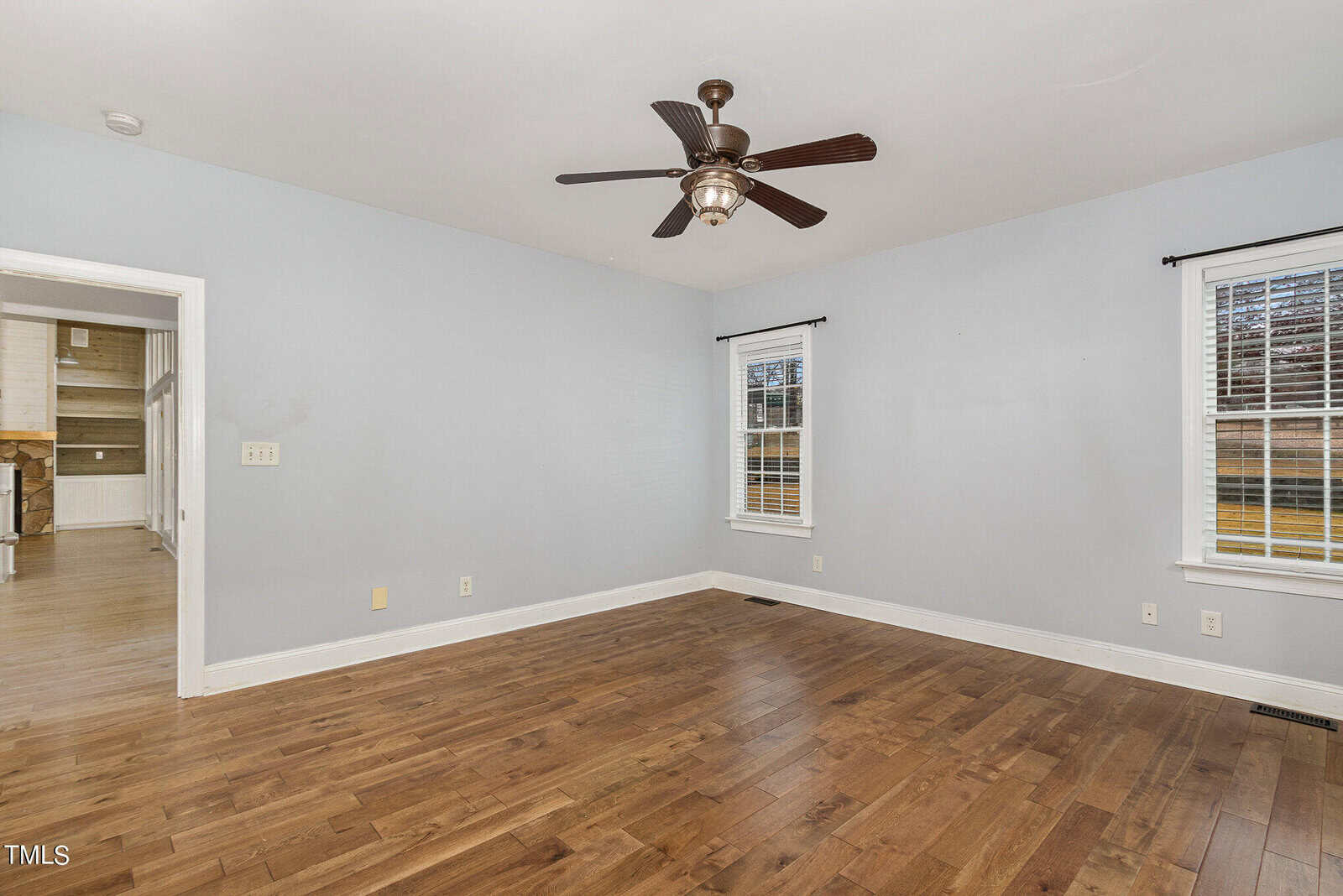 An empty room with light gray walls, two windows with white blinds, and a ceiling fan. The floor is wooden, and a doorway leads to another room with cabinetry. Natural light enters through the windows, illuminating the space.