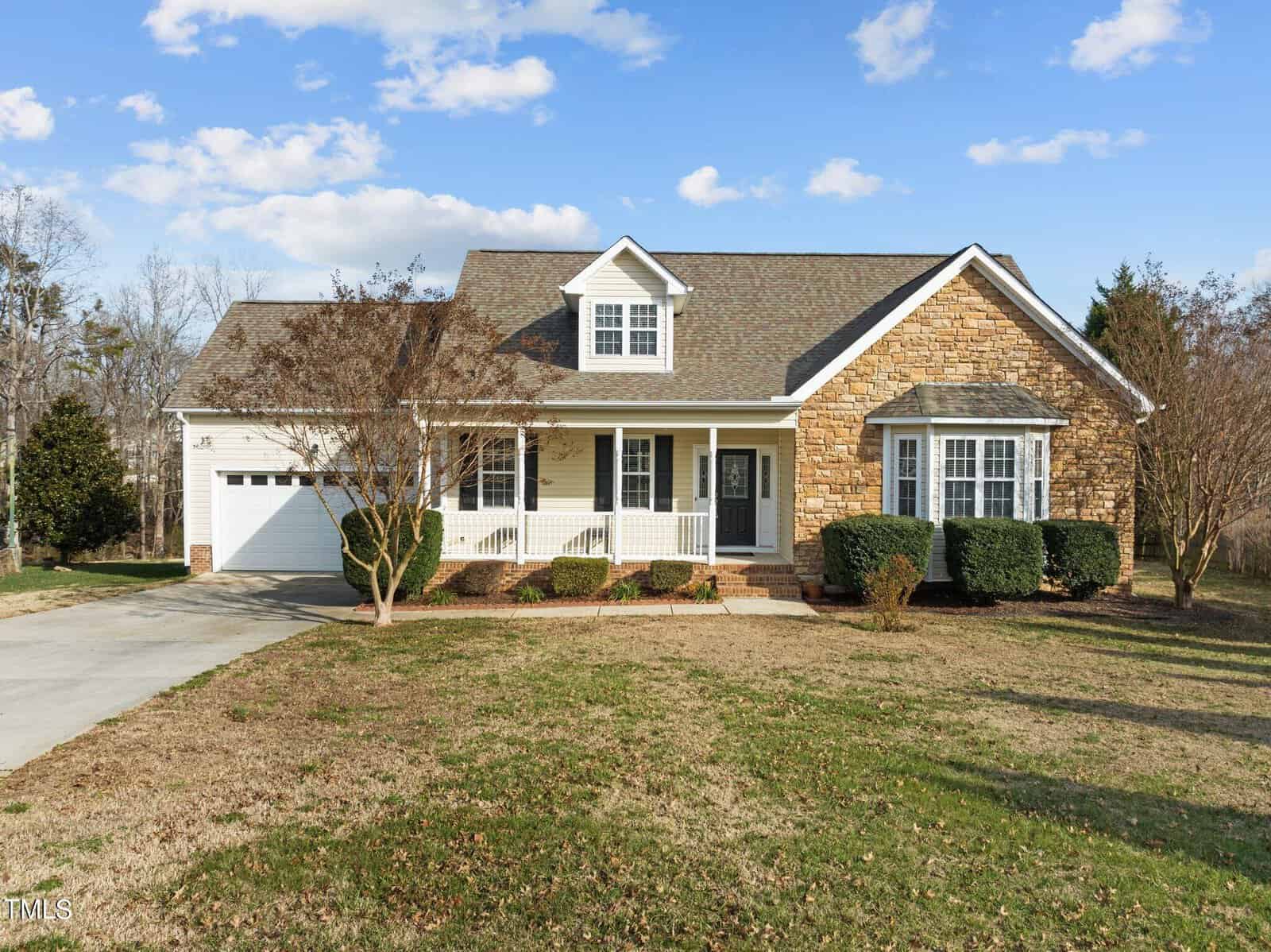The single-story house at 3527 Bluebonnet Drive in Wake Forest boasts a charming stone and siding exterior with a gabled roof. A covered front porch with white railings is framed by shrubs, and the driveway leads to an attached garage. The sky above is partly cloudy, completing this picturesque setting.