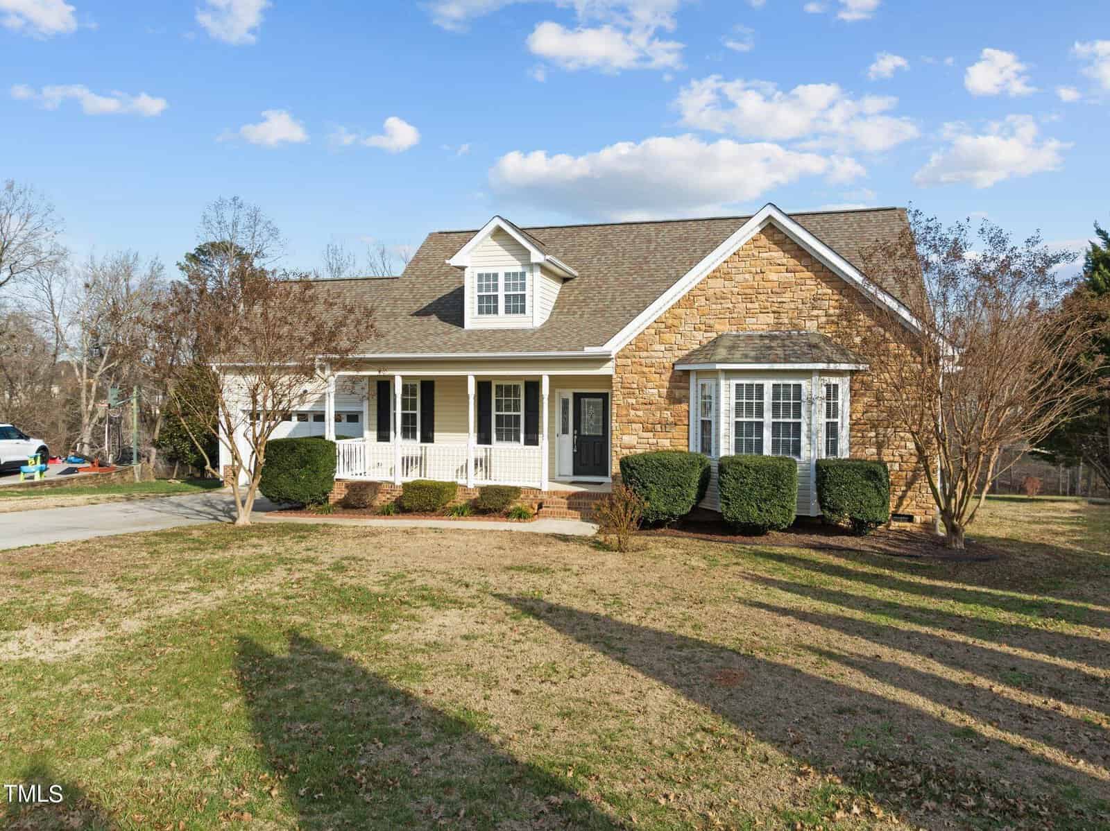 A single-story house with a stone and siding exterior, a covered front porch, and a neatly trimmed lawn. It features a large bay window, a paved driveway, and is surrounded by leafless trees under a partly cloudy sky.