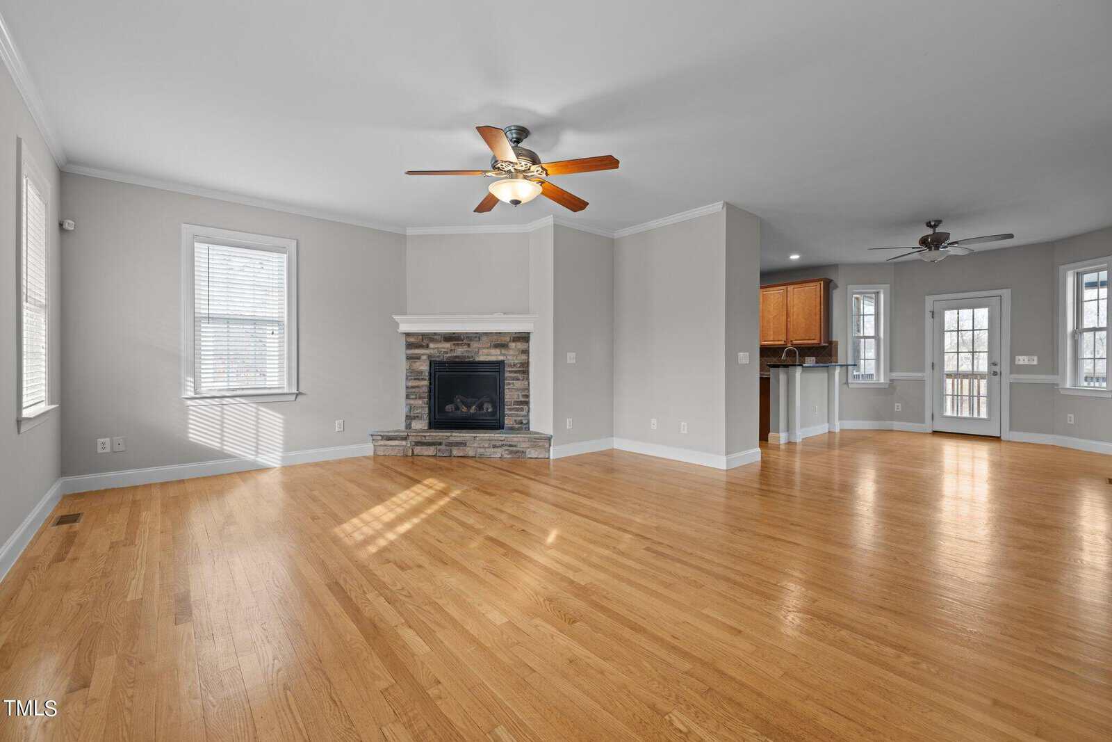 Spacious living area with hardwood floors, light gray walls, and large windows. Features a corner stone fireplace and ceiling fan. Open floor plan leads to a dining area and a partial view of the kitchen at the back.