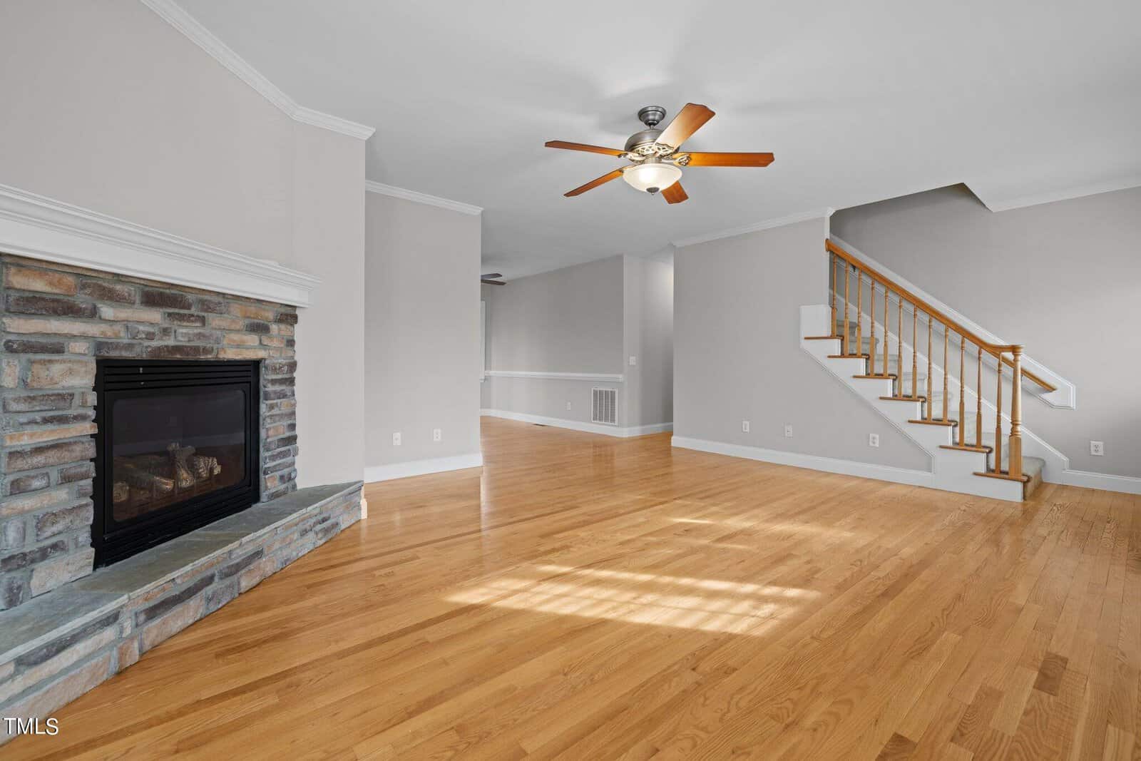 A spacious living room with light wood flooring and pale gray walls. A stone fireplace is on the left, and a wooden staircase with white railing is on the right. A ceiling fan with wooden blades hangs from the ceiling.