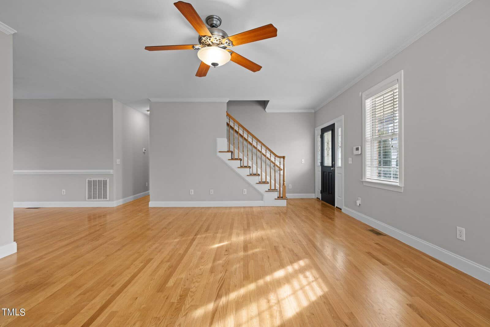 Spacious living room with light hardwood floors and gray walls. A ceiling fan with wooden blades is centered overhead. A staircase with a wooden banister is visible, and large windows let in natural light.