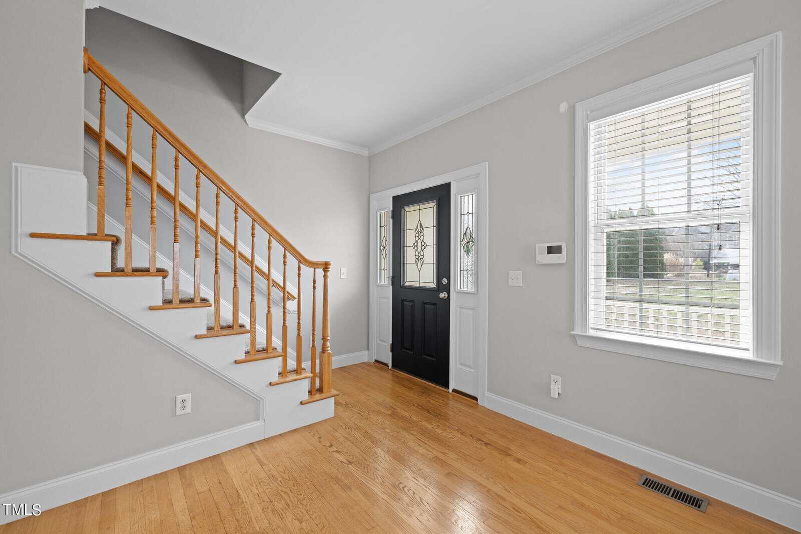 A bright, empty foyer with light wood flooring and soft gray walls. A wooden staircase with a natural finish is on the left. The black front door, flanked by decorative glass panels, is centered. A window with blinds is on the right.