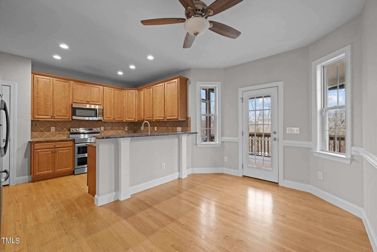 A spacious kitchen with wooden floors, light green walls, and recessed lighting. It features wooden cabinets, stainless steel appliances, a central island with a bar top, and a ceiling fan. A door and windows open to a deck outside.