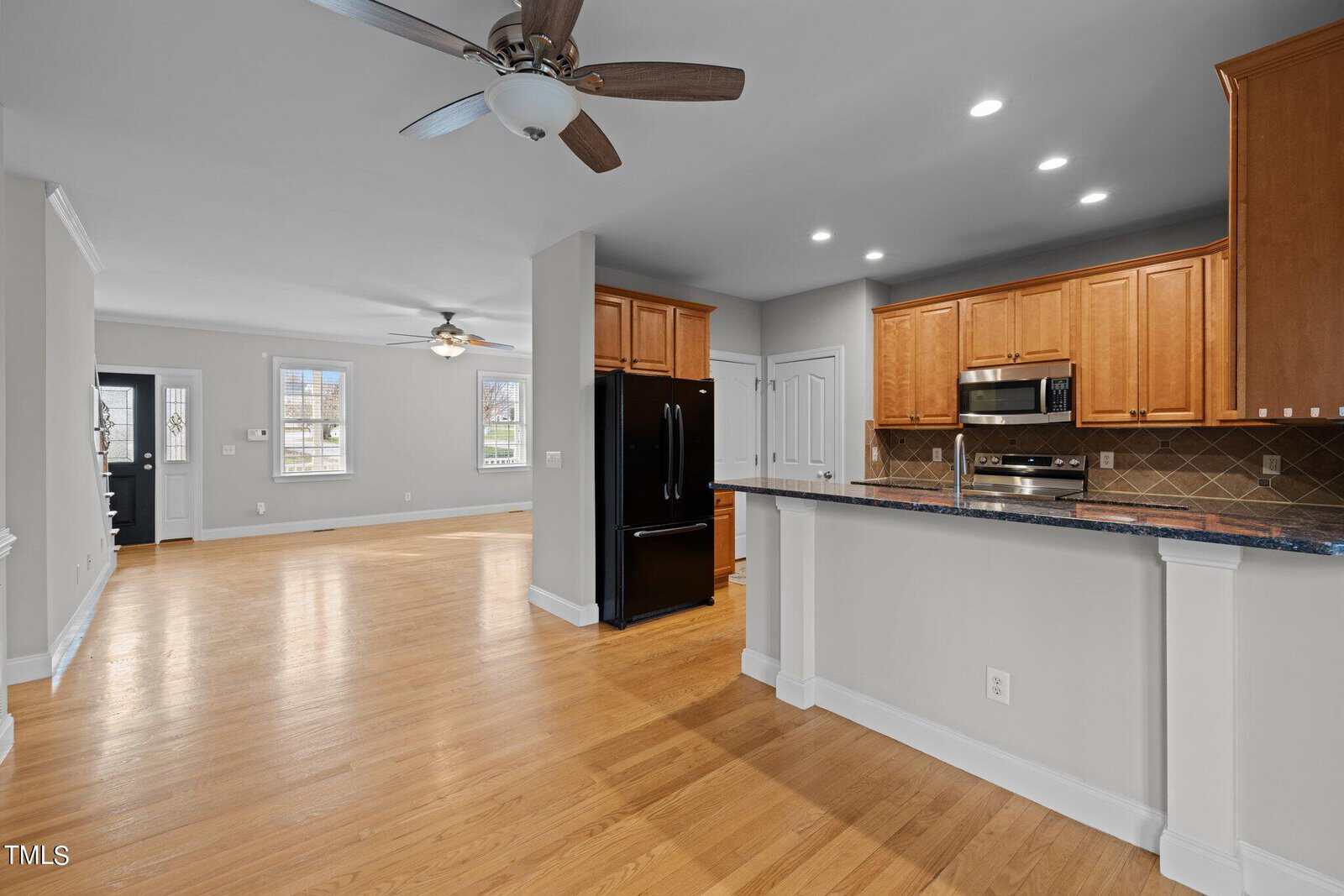 Spacious open-plan kitchen and living area featuring hardwood floors, light gray walls, a ceiling fan, wooden cabinets, and stainless steel appliances. Large windows provide natural light.