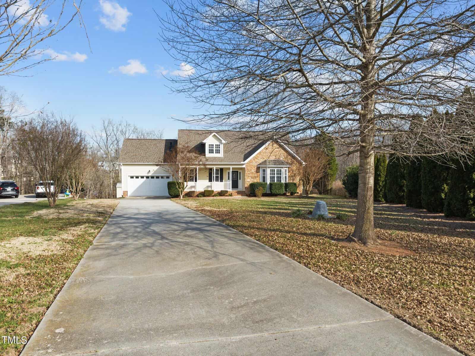 Suburban house with a sloped roof and a front porch, surrounded by a grassy lawn and trees. A concrete driveway leads to a garage. The sky is clear and sunny.