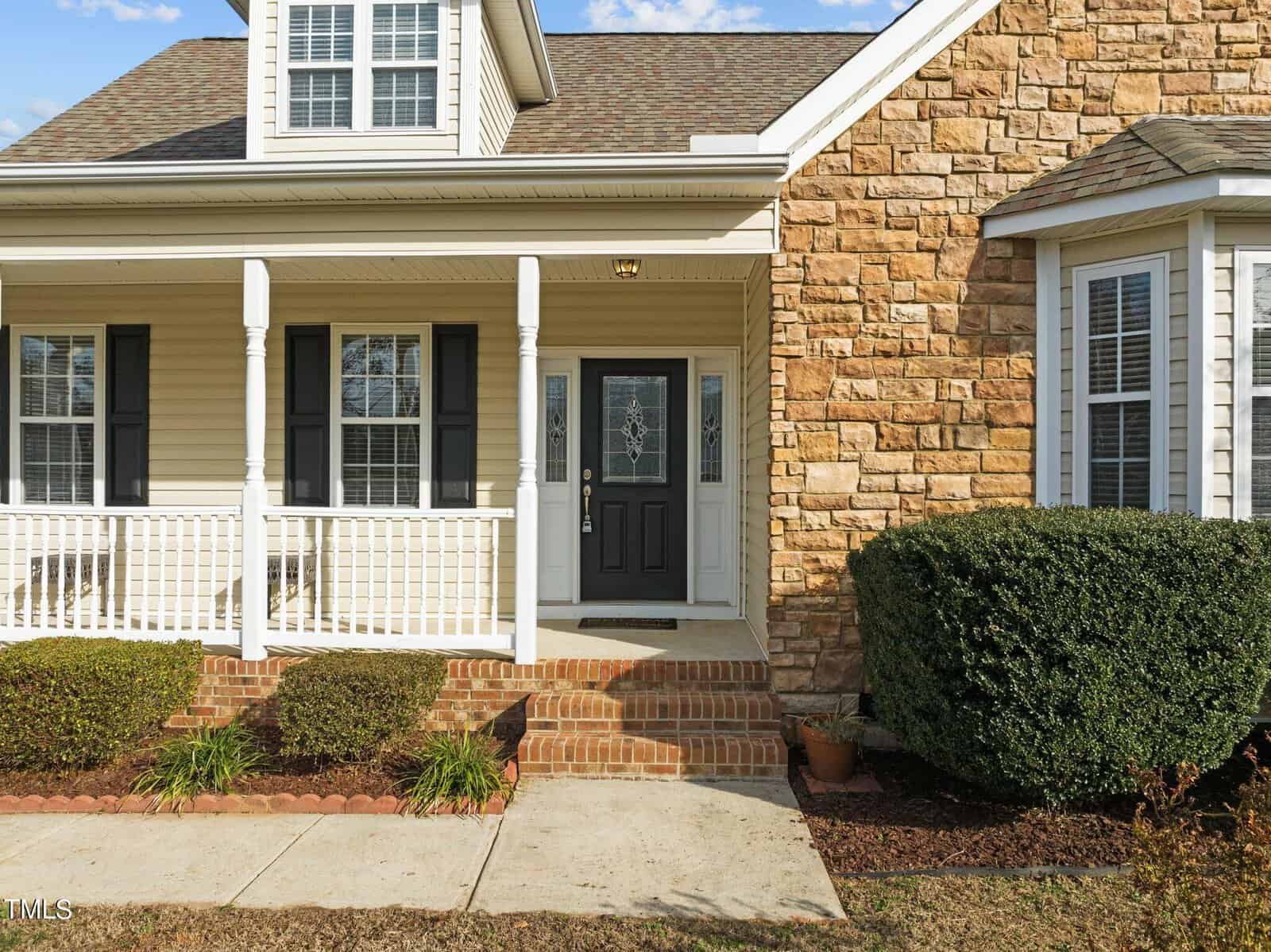 A two-story house with light-colored siding and stone accents. It features a covered front porch with white railings and columns, a black front door with decorative glass, and manicured shrubs lining the brick walkway.