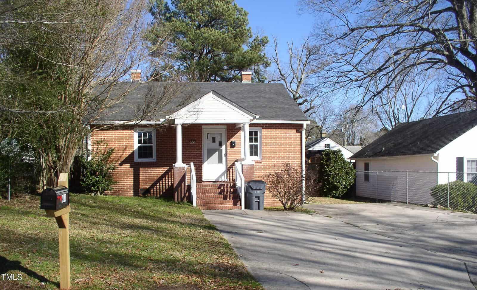 A red brick, single-story house at 408 Oak Street, Henderson NC, with white trim and a central white door. It features a driveway, mailbox, leafless trees, and a detached garage