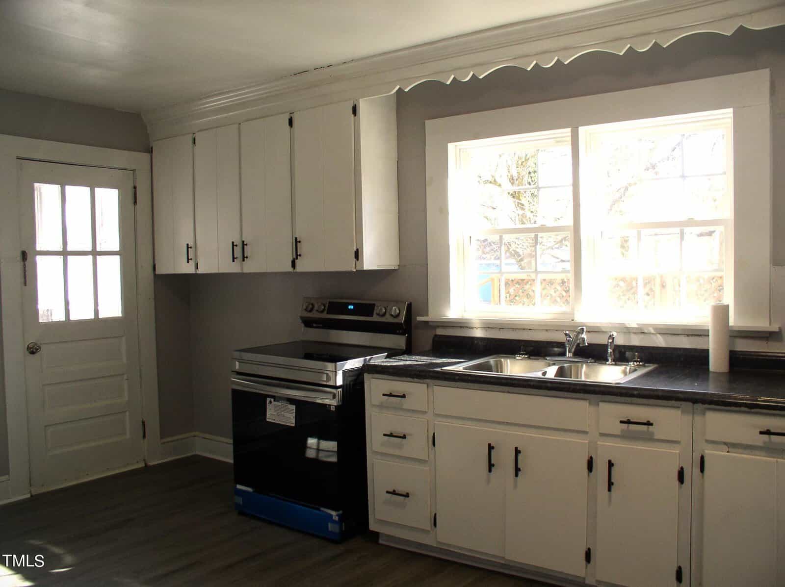 A bright, spacious kitchen with white wooden cabinets, black countertops, modern appliances, a large window, and a white door. empty and clean with a scalloped valance above the sink.