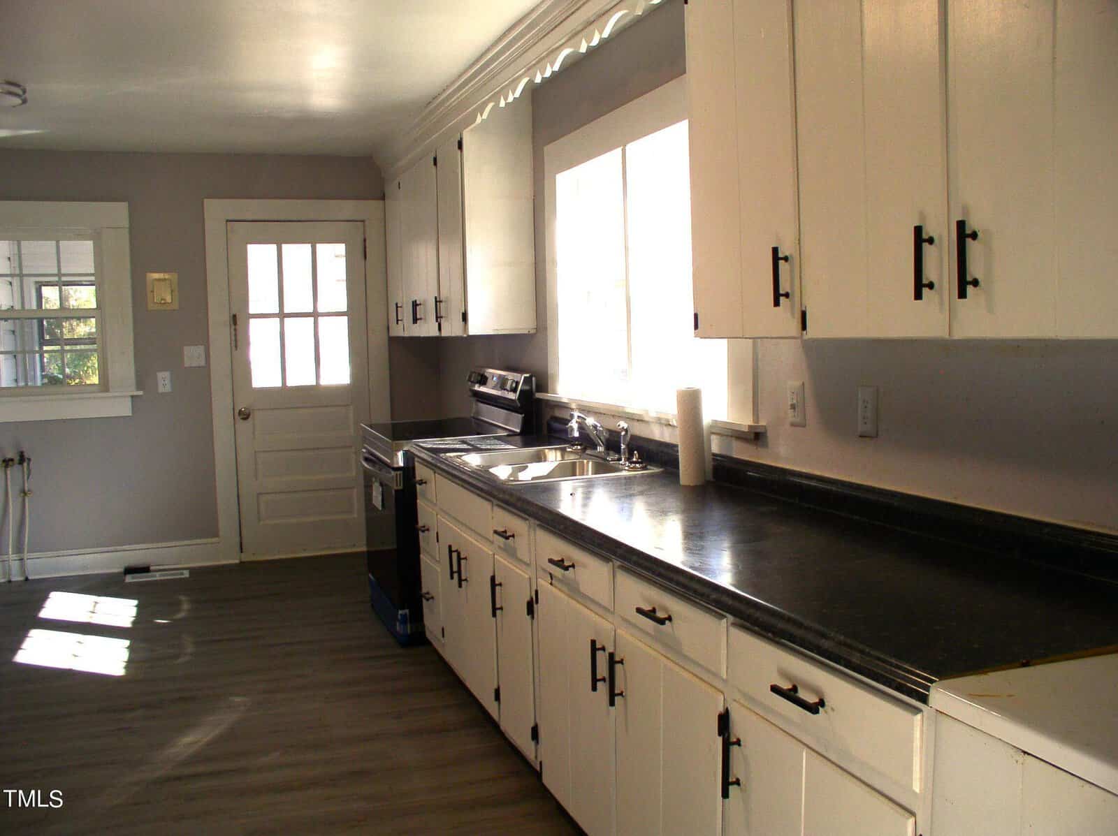 A bright, clean kitchen with white cabinetry, dark countertops, and wooden flooring. the room has a sink, a window with blinds, and multiple cabinets, with sunlight streaming in.