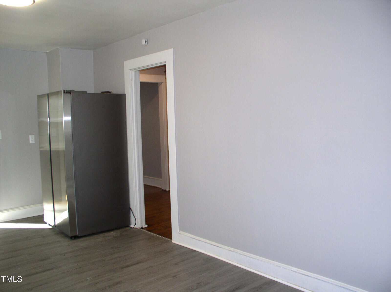 Empty kitchen with gray flooring and light gray walls, featuring a stainless steel refrigerator, and an open door leading to another room.