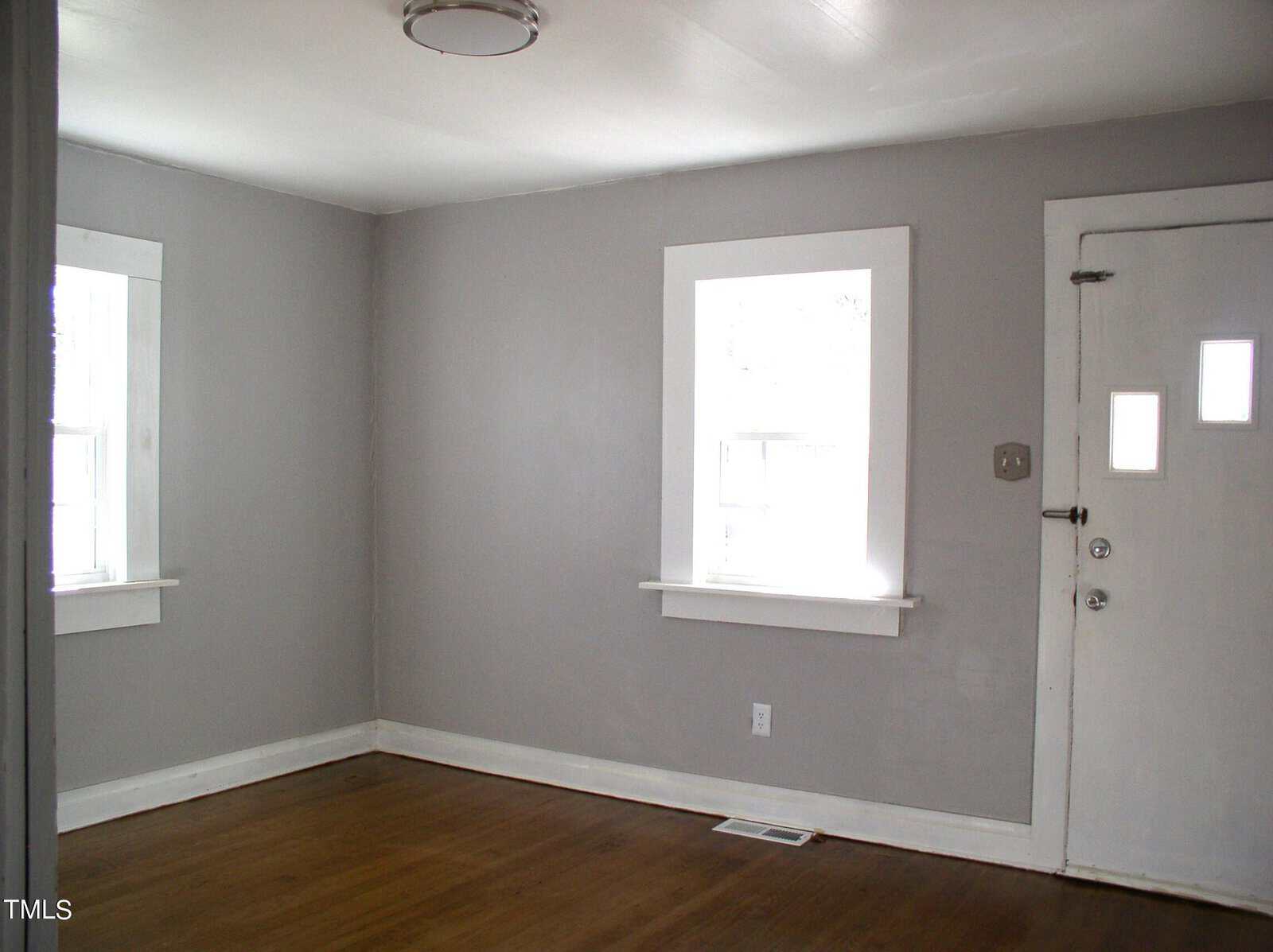 An empty room painted in light grey with hardwood flooring, featuring two windows with white trim and a closed white door.