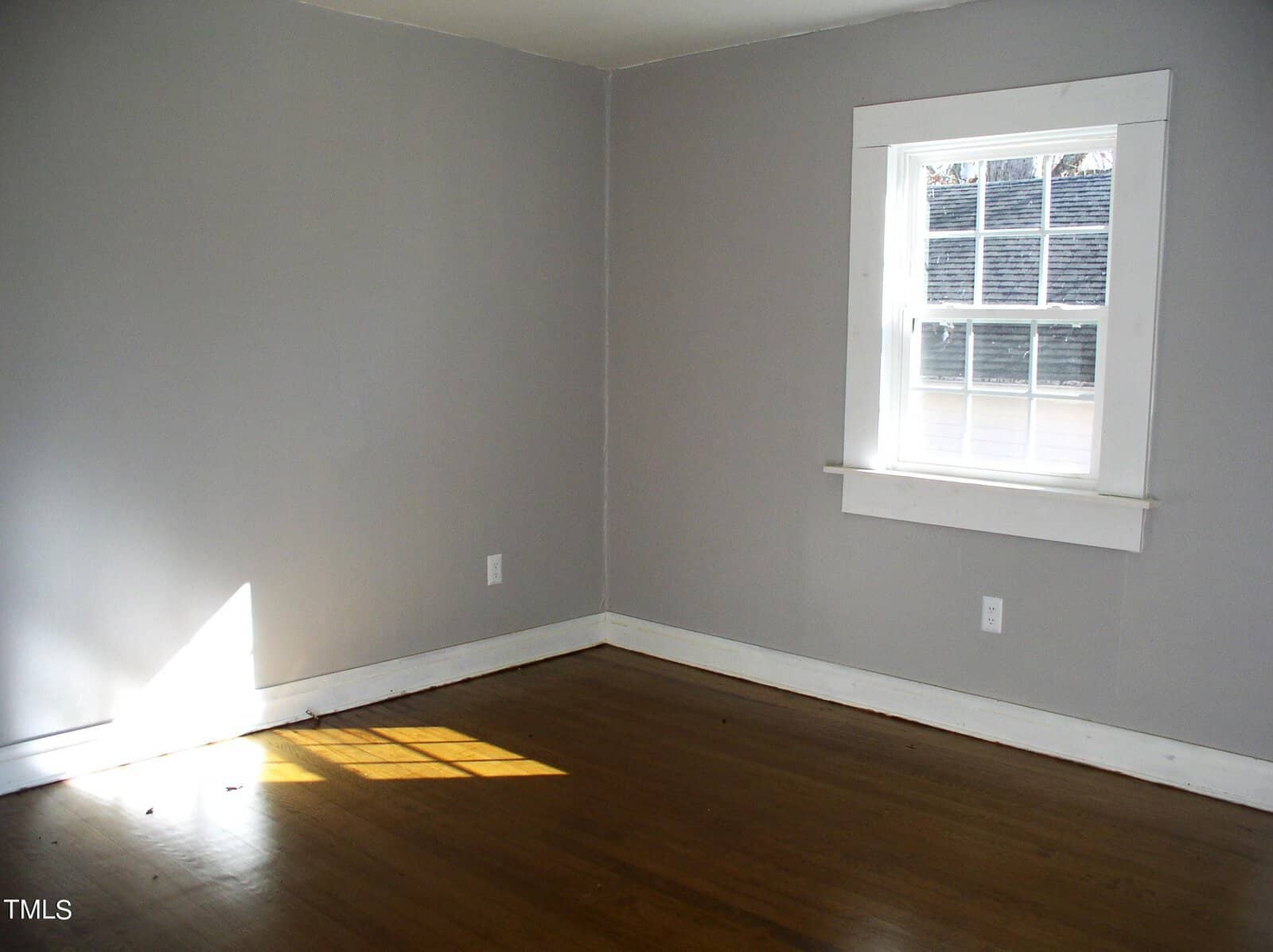 Empty room with light grey walls and hardwood floor, featuring a single white-framed window letting in sunlight.