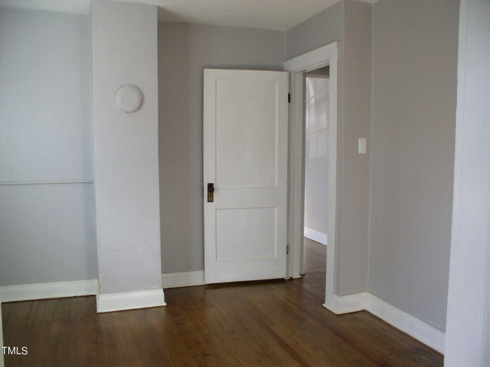 An empty room with light gray walls and hardwood flooring. there's a closed white door in the center with a round decorative element on the wall to its left.
