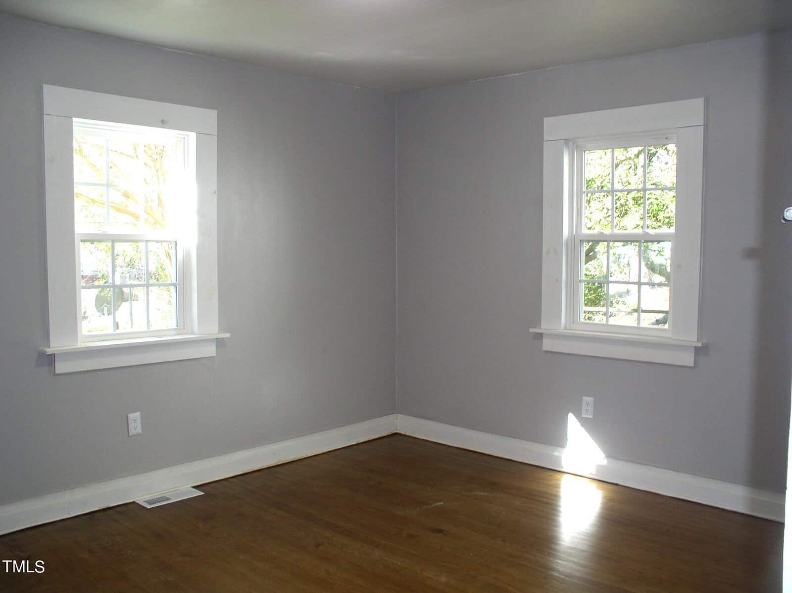 Empty room with light gray walls and hardwood floor, featuring two windows with white trim that provide a view of green foliage outside.