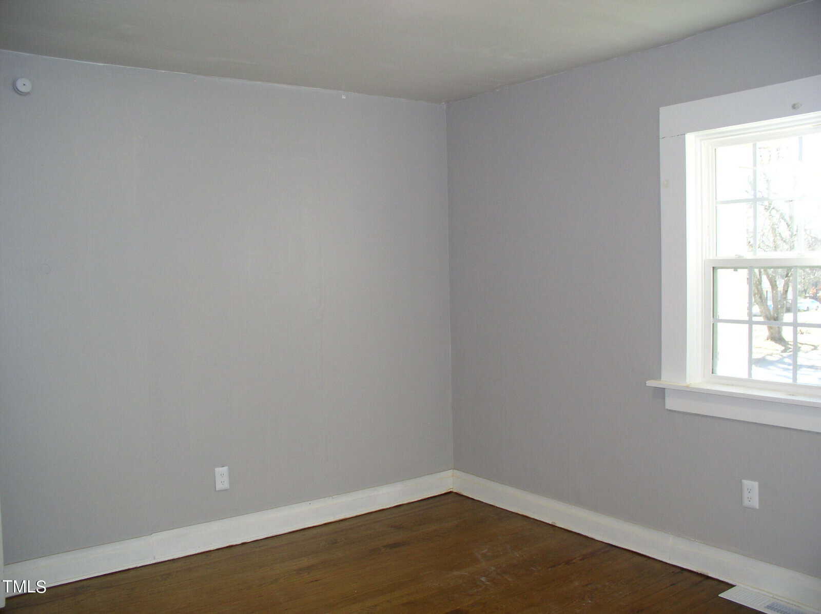 Empty room with gray walls and a hardwood floor. one window on the right side lets in natural light. electrical outlets visible on the walls.