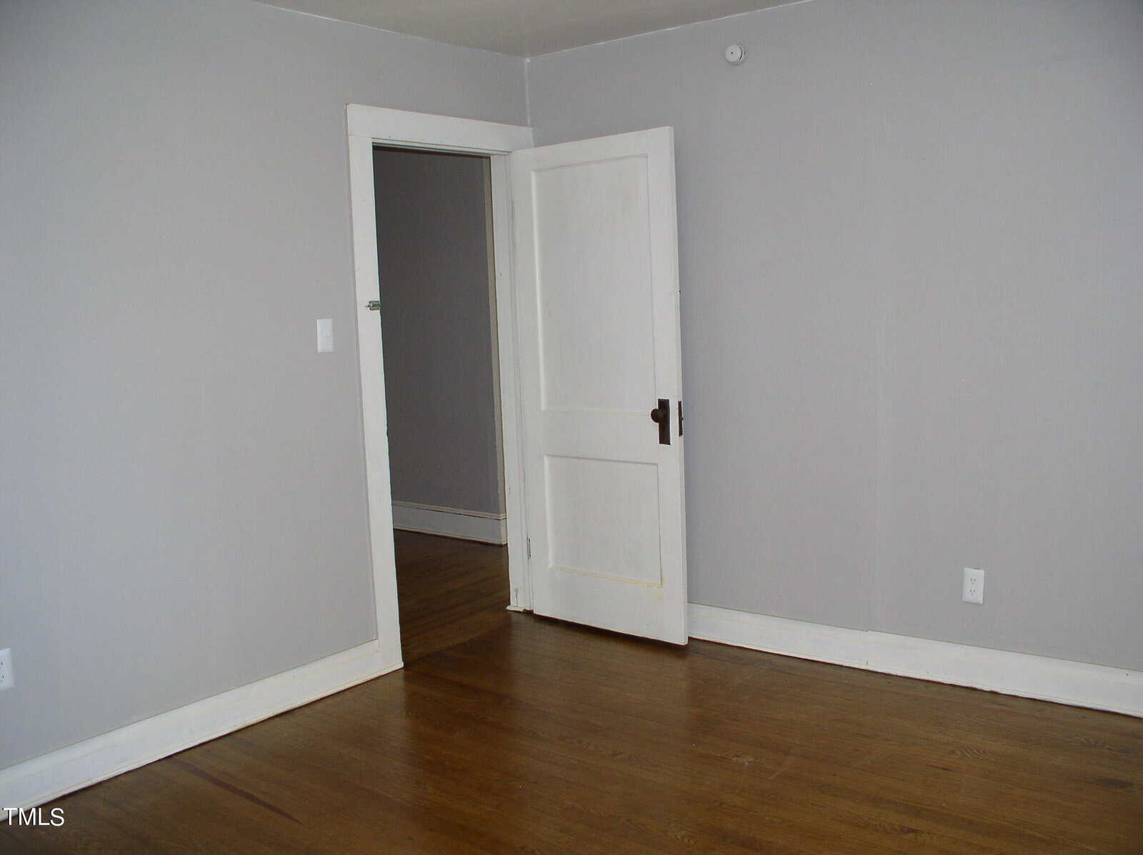 Empty room with gray walls and hardwood floor featuring an open white door at the center, leading to another room.