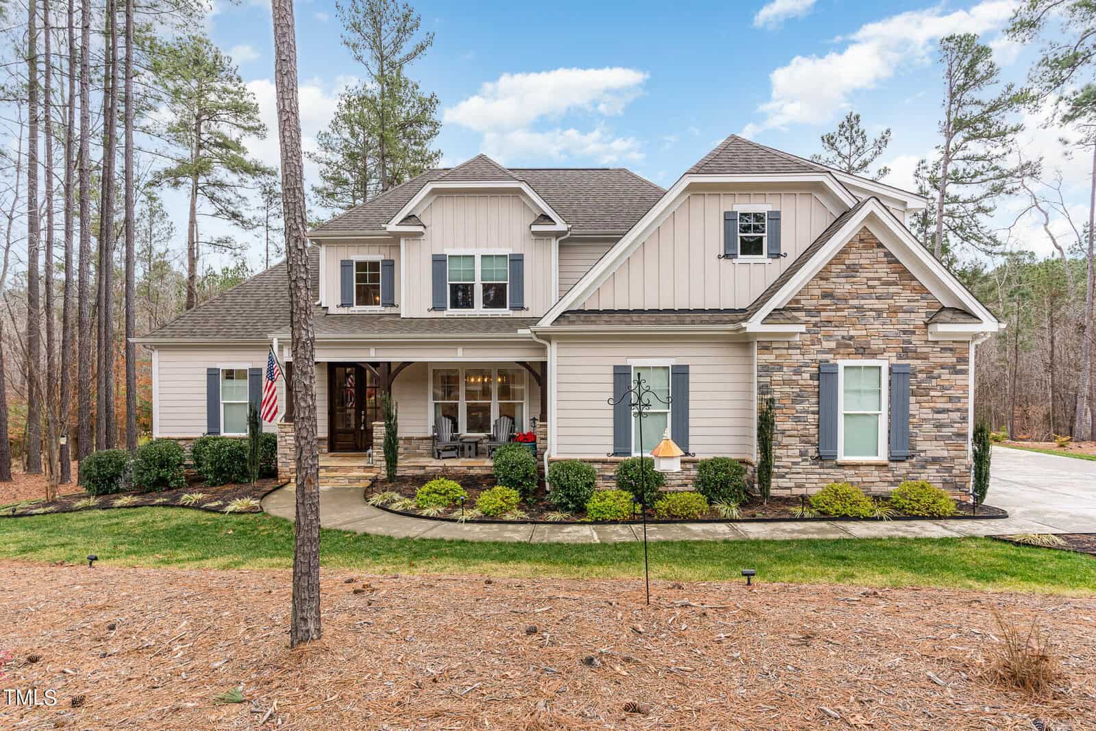 A large, modern two-story house at 3597 Carole Court, Wake Forest, boasts stone and beige siding surrounded by a landscaped yard. An American flag proudly flutters near the front porch adorned with red chairs. Tall trees and a partly cloudy sky serve as the serene backdrop.