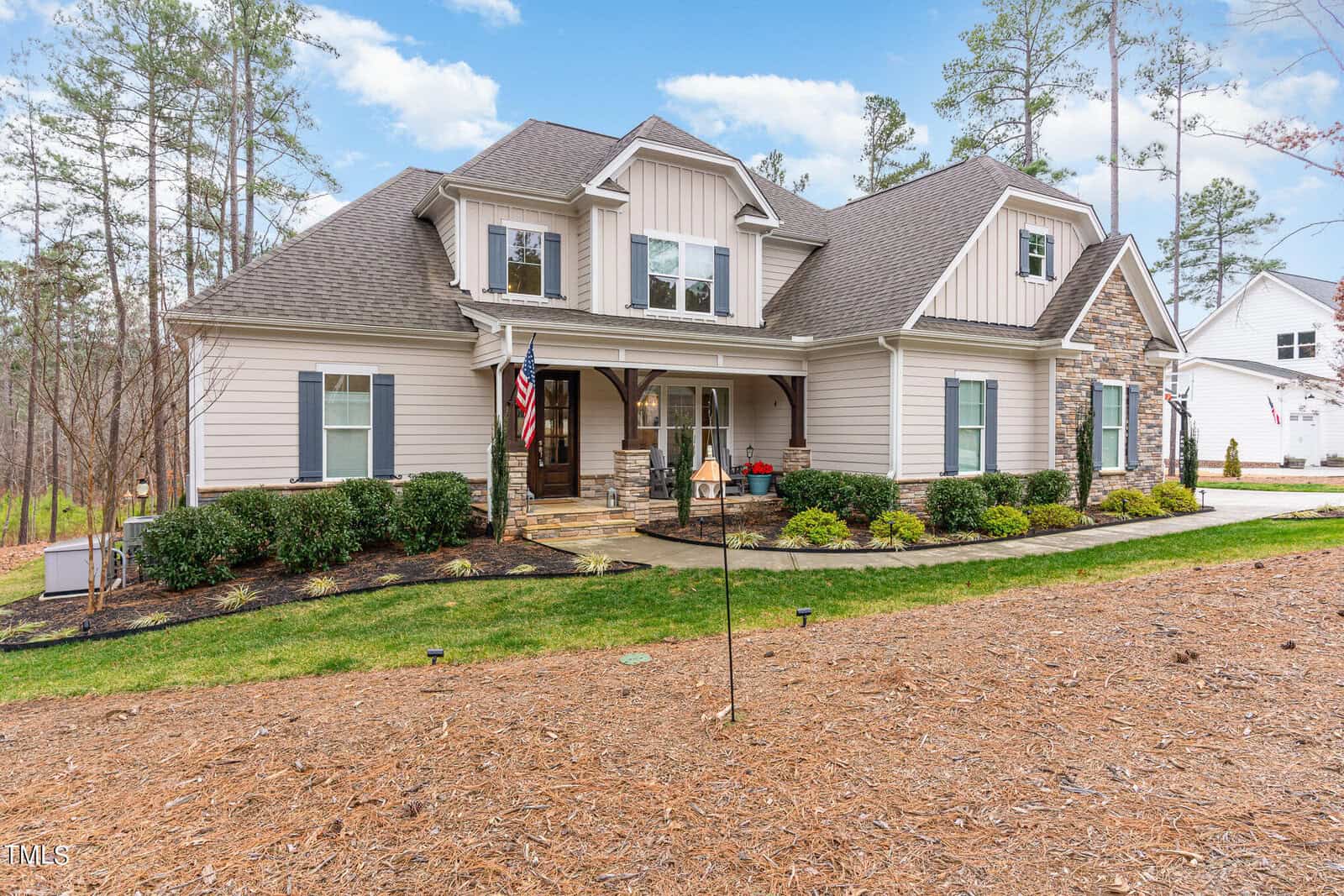 A modern two-story house with beige siding and stone accents, featuring a covered front porch and American flag. The front yard has manicured bushes and a well-maintained lawn, surrounded by tall trees under a partly cloudy sky.